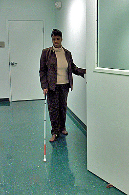 Two photos show a woman walking along a hall, approaching the edge of an open door.  Her left hand trails the wall and her right hand holds a cane with the tip on the floor straight ahead of her hand.  The cane never touches the door, and she is about to collide into it.